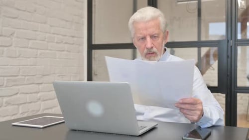Senior Man Reviews Documents at Desk with Laptop