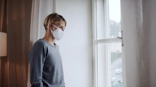 Woman in Mask Looks Out Window at Home