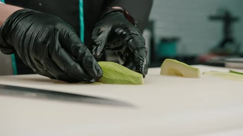 Close-up of a sushi maker in gloves cutting avocado with a professional kitchen knife on a board