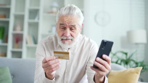Happy elderly senior man doing online shopping typing credit card number on smartphone sitting