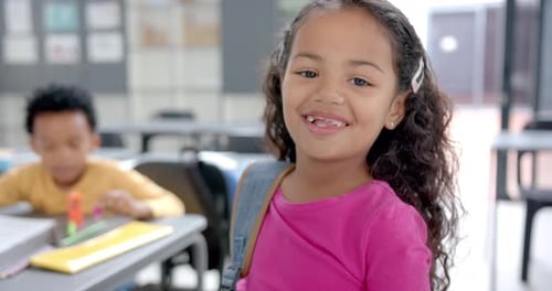 In school, smiling girl with backpack standing in classroom, looking at camera