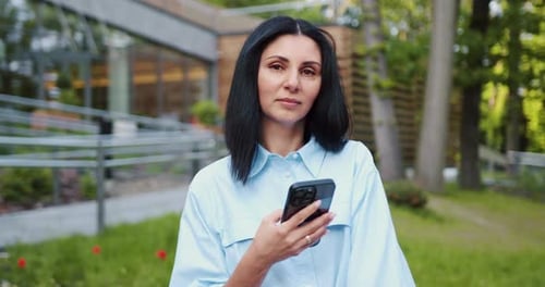 Portrait of Caucasian woman with dark hair using mobile phone with a pleased smile, standing in fron