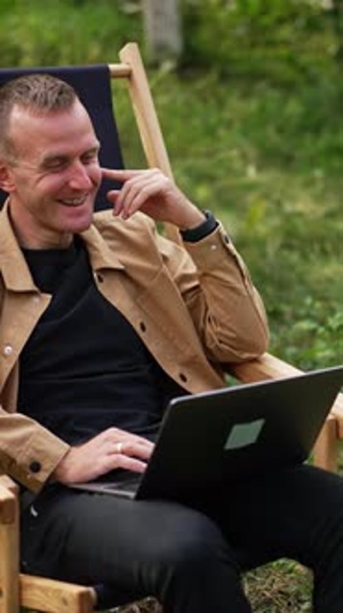 Caucasian man in brown shirt sitting with laptop on his laps in the garden.