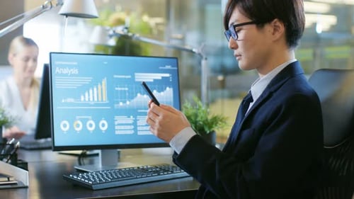 In the Office Asian Businessman Uses Smartphone, Typing Important Message. His Desktop Computer Sho