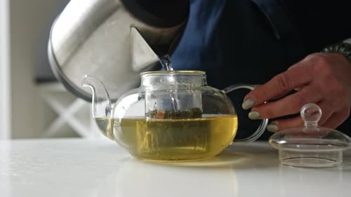 Girl Pouring Hot Water From The Kettle Into Teapot With Green Tea In The Kitchen