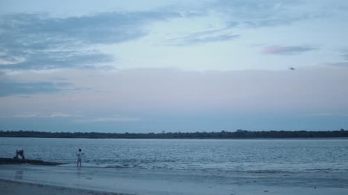 Two elderly people stand on the shore of a reflective beach fishing under the fluffy, white clouds f