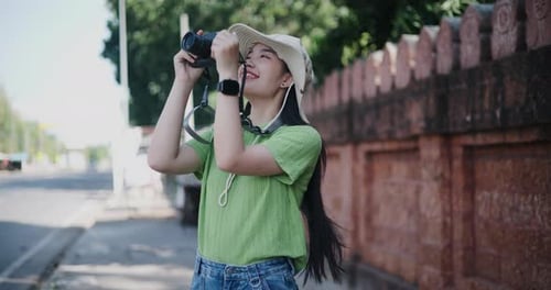 A young Asian woman stands on the side of the road and enjoys taking pictures with a camera.