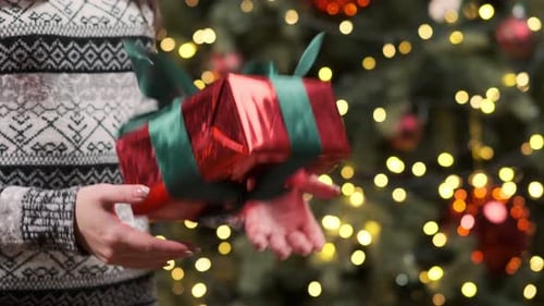 Close-up of a girl in a Christmas sweater holding and tossing a red gift box with a green ribbon. Be
