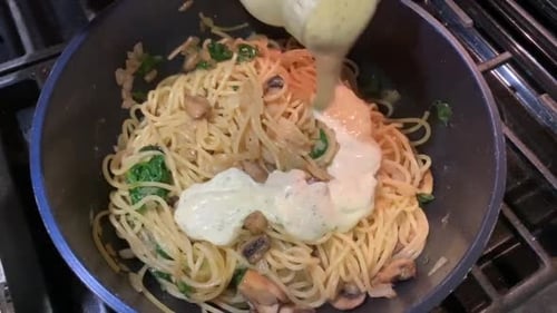 Pouring a jar of white and creamy Alfredo sauce over cooked pasta noodles with sauteed vegetables. O