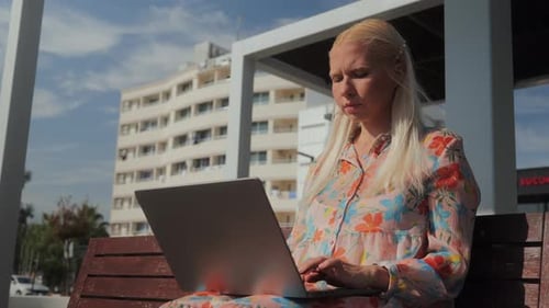 Young Beautiful Blonde Woman is Sitting on a Park Bench and Working on a Laptop
