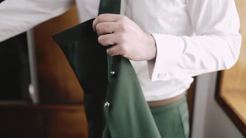 Man in a white shirt and green tie holding a green jacket, preparing for a formal event.