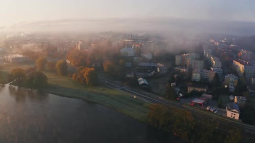 An aerial view of a misty town at sunrise, fog hovering over buildings and a river winding through t
