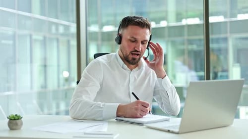 Businessman in White Shirt with Headset in Office Video Call Taking Notes on Laptop