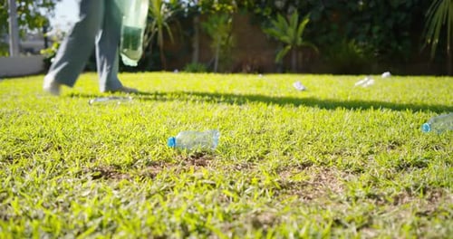 Botella detectando, mujer con guantes caminando por el césped agachándose y recogiendo basura en una bolsa verde