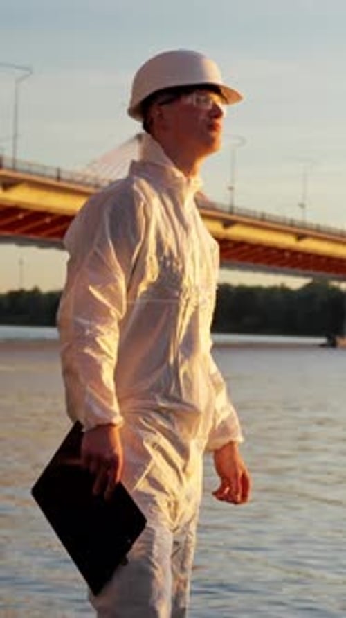 Young caucasian environment inspector in a hazmat suit and hard hat standing by a river and bridge