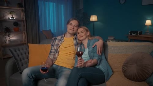 A Young Couple Sits Hugging on the Couch with Glasses of Red Wine and Watching TV A Man and a Woman