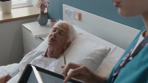 Nurse Using Tablet and Listening to Senior Female Patient Lying in Hospital Bed