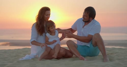 A Married Couple with Their Daughter Sitting on the Ocean Shore During Golden Hours Enjoys Time