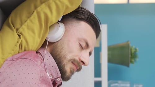Vertical Video of Man relaxing on sofa at home with headphones listening to music, daydreaming.