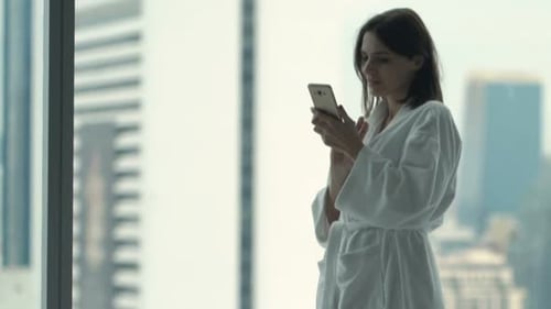 Woman in bathrobe using phone by city view window
