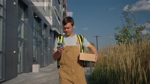 Delivery Courier in Overalls Walking Through a Modern City Street Holding a Parcel and Checking a