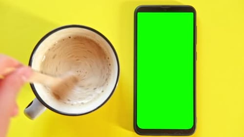 Smart Phone with Empty Green Screen Next to Coffee Cup Person Hand Stirring Sugar with Spoon Against