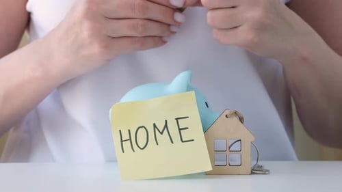 Woman Putting Coins Into Piggy Bank with Paper Label Home Next to House Keyring