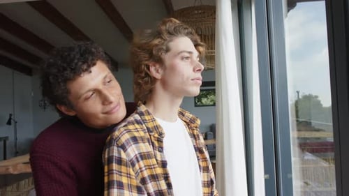 Happy diverse gay male couple looking through window and embracing at home, slow motion