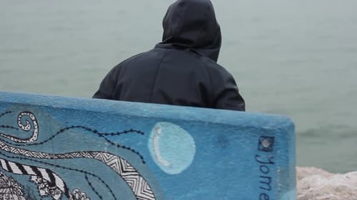 Hooded Figure Sits on Bench Overlooking the Ocean