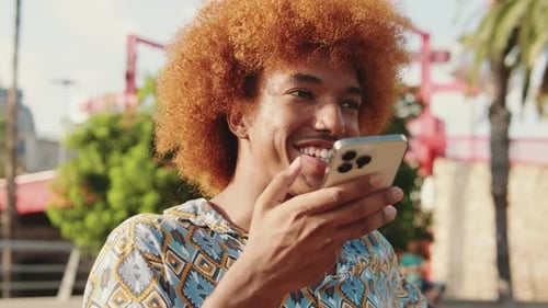 Close-up of young guy sitting on bench, chating online with smartphone in his hands and smiling