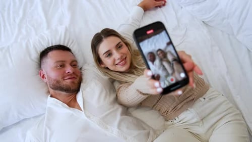 Ein
glückliches kaukasisches Paar liegt auf dem Bett und macht ein Selfie. Verliebte küssen sich liebevoll. Ansicht von oben.