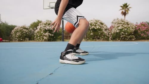 Young Athlete Dribbles With Basketball Under His Legs In Outdoor Field
