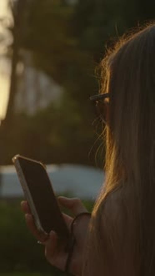 Vertical video. A long-haired brunette looks at her phone in the sunset, view from behind.