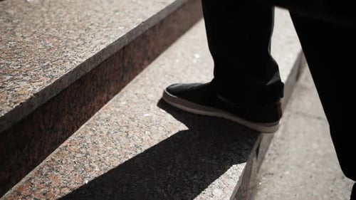The Businessman's Feet Go Up the Stone Stairs Slow Motion Sunny Day Black Pants and Boots of a