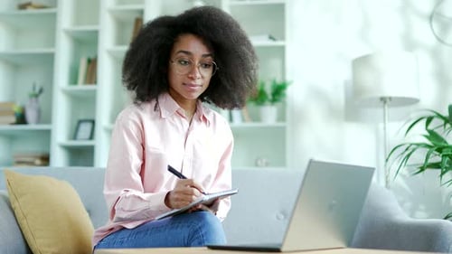 Woman Writes in Notebook While Looking at Laptop