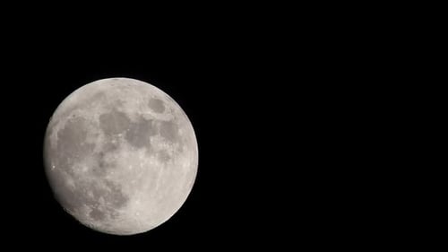 Bright full moon set against dark black sky, moving from bottom left of frame towards center left of