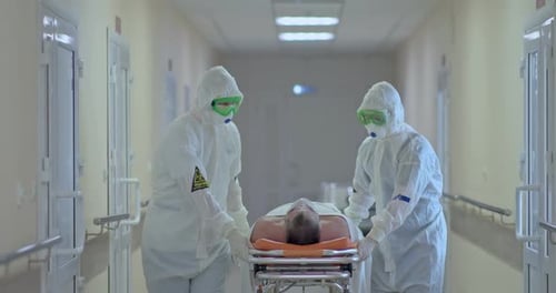Doctors in protective suits carry a sick patient on a stretcher along the hospital corridor.