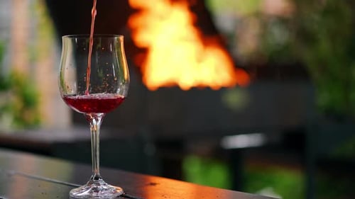Closeup Glass on the Left at Background of Bbq Fire and Red Wine Pouring in Slow Motion Closeup