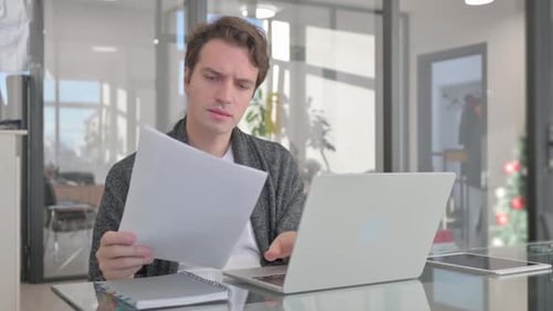 Man Reviews Documents and Types on Laptop in Office
