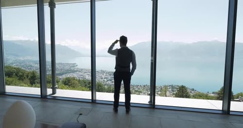 A Man Near a Panoramic Window Enjoying View of a Serene Lake and Majestic Mountains