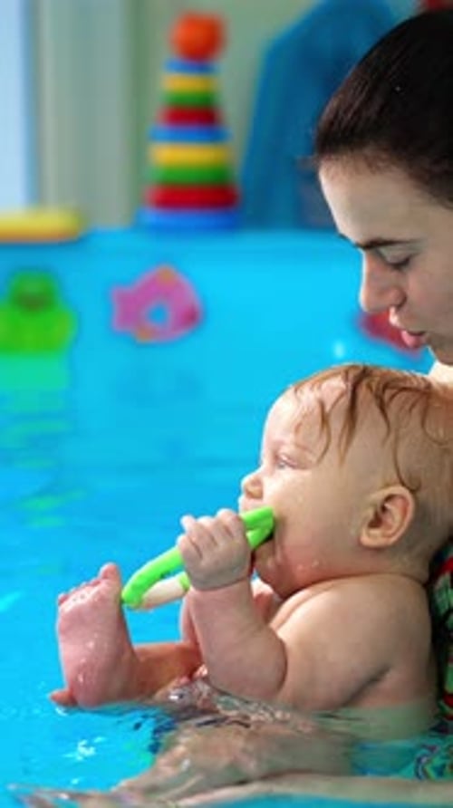 Woman with a little baby boy in the swimming pool. Adorable infant holds the toy in his mouth.