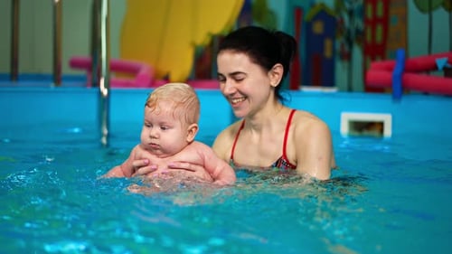 Adorable chubby baby is in the swimming-pool. Brunette woman teaching the infant to swim.