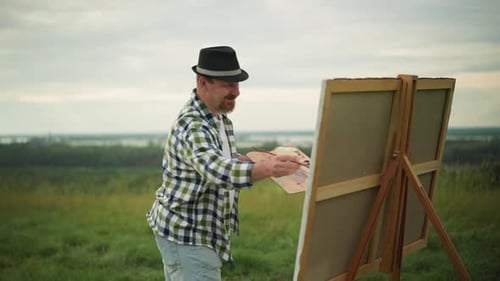 Focused Painter in Hat and Checkered Shirt Working on Canvas in a Grass Field