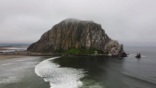 Aerial zoom out shot of waves rolling onto the beach and Morro Rock in coastal California. The rock