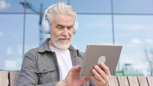 Aged Gentleman Using Tablet and Headphones in Megapolis