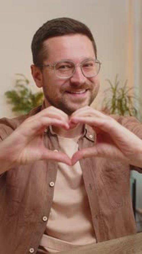 Happy Caucasian Young Man Makes Symbol of Love Showing Heart Sign to Camera at Home Office Desk