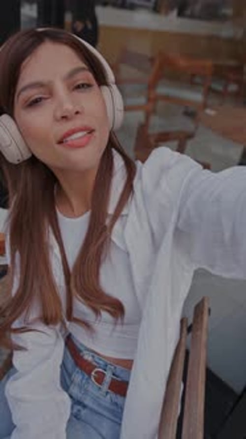 Young Woman Taking a Selfie Video with Headphones at a Sidewalk Cafe