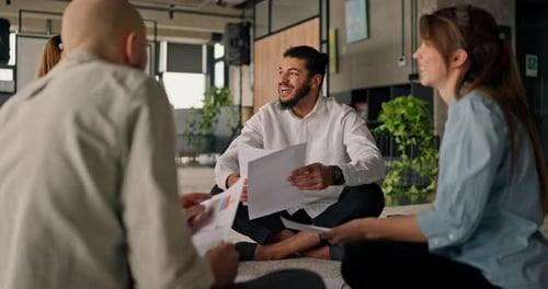 A Happy African American Office Worker Chats with Colleagues While Sitting on a Carpet Reviewing