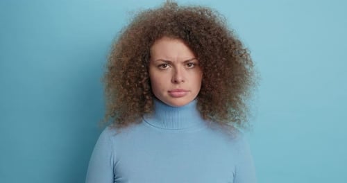 Dissatisfied european woman with curly hair smirks in studio portrait on blue