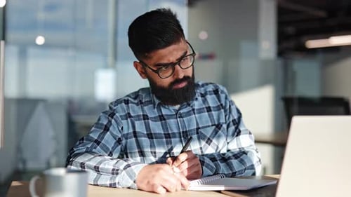 Young Businessman Writing in Notebook at Office Desk with Sunlight